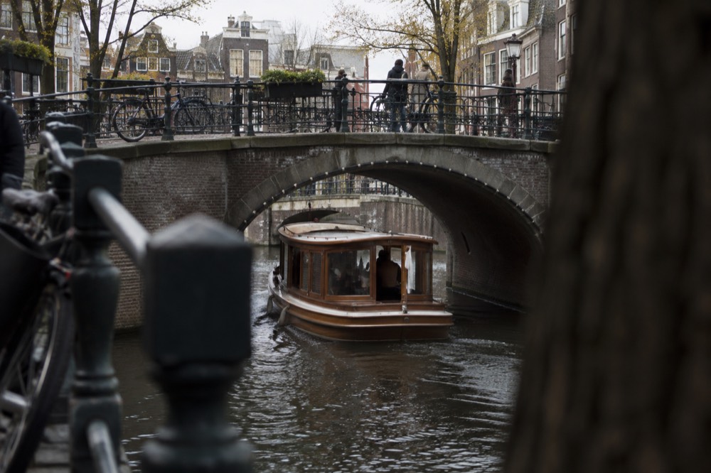 A wooden saloon boat passing under a low arched canal bridge in Amsterdam