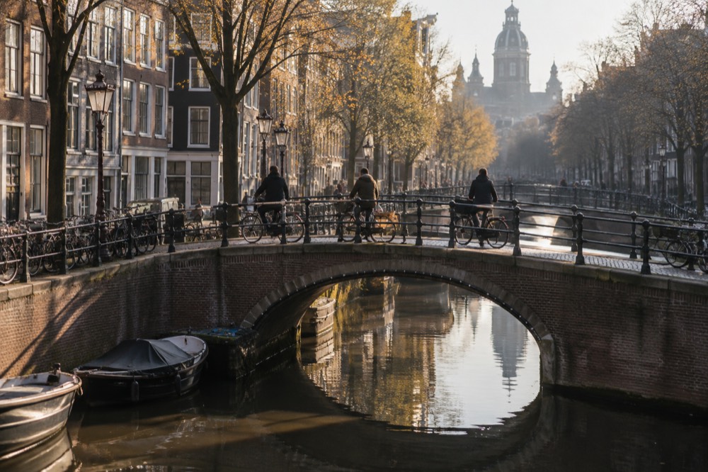 Cyclists crossing a small arched bridge over an Amsterdam canal lined with narrow brick houses
