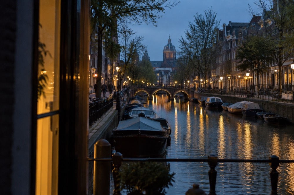A narrow Amsterdam canal at dusk with lit bridges and moored boats