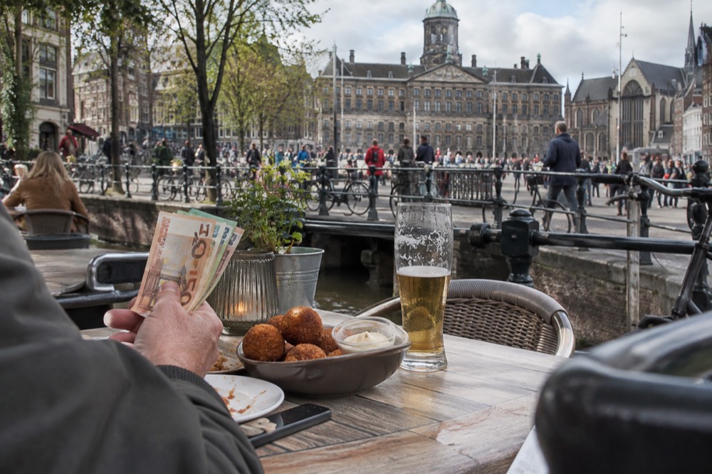 A traveller's hand holding a stack of euro notes beside a bowl of bitterballen on an Amsterdam canal-side terrace