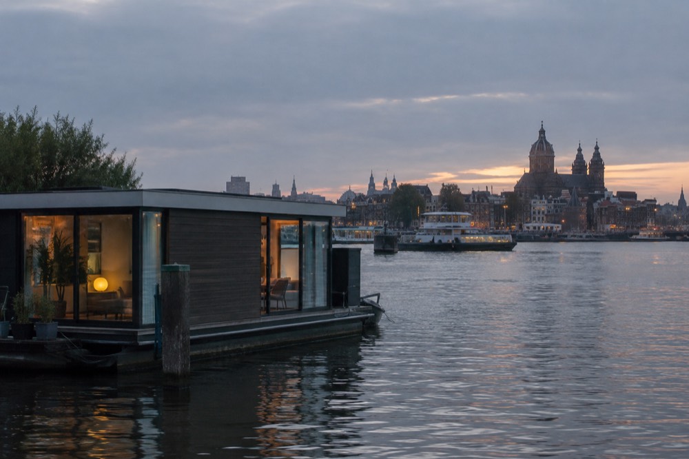 A modern houseboat on the IJ waterfront with the Amsterdam skyline across the water at dusk