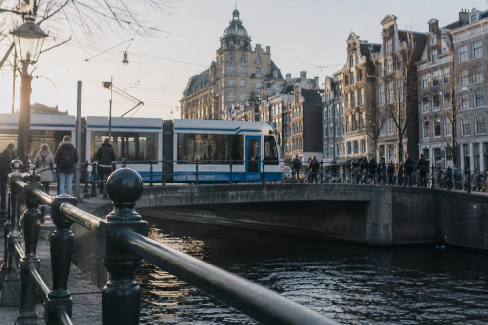 A tram passing a canal bridge with pedestrians waiting on the sidewalk