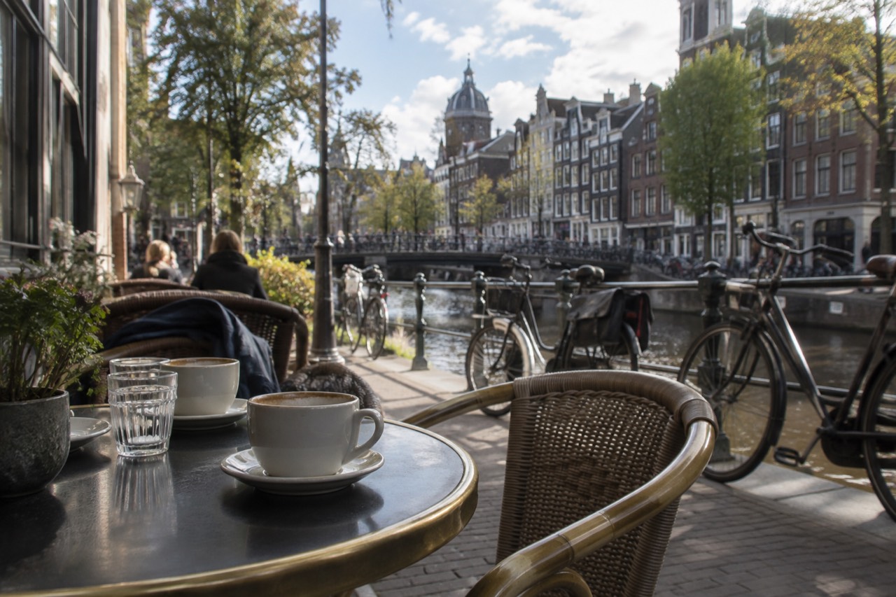 A canal-side café terrace in Amsterdam with small tables and bicycles parked along the railing