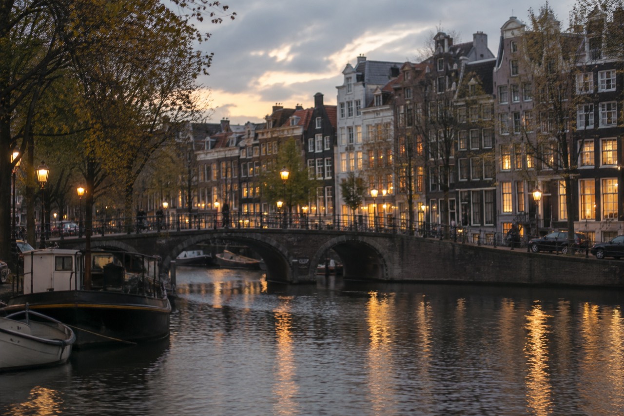 A canal bridge in Amsterdam at dusk with lit gabled houses reflected in the water