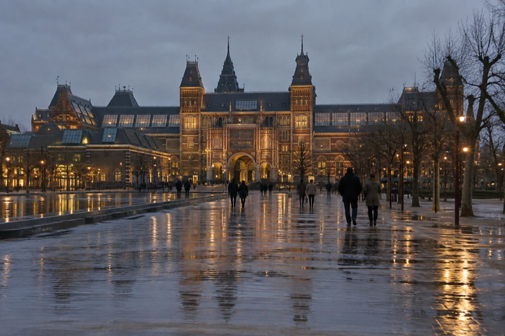 The illuminated Rijksmuseum facade and Museumplein at night with wet pavement reflecting the lights