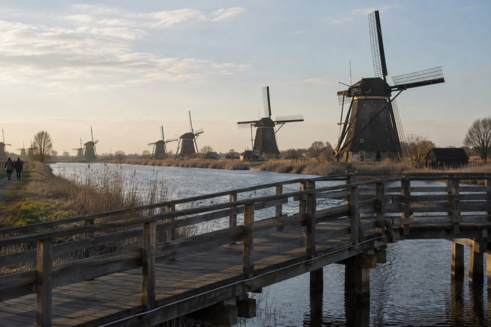 A row of traditional Dutch windmills standing beside a river with a wooden footbridge in the foreground