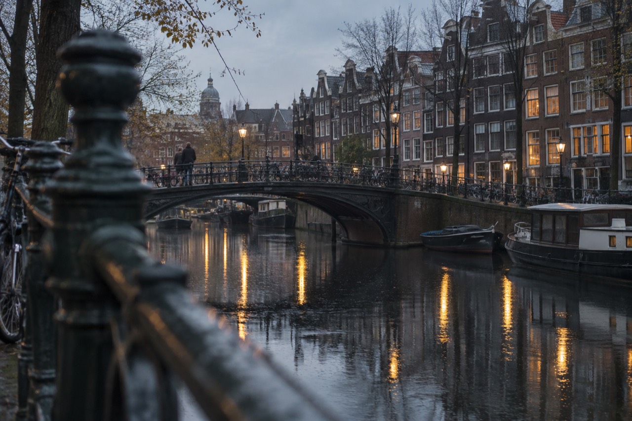 Amsterdam canal houses reflected in still water at dusk with a curved iron bridge in the foreground