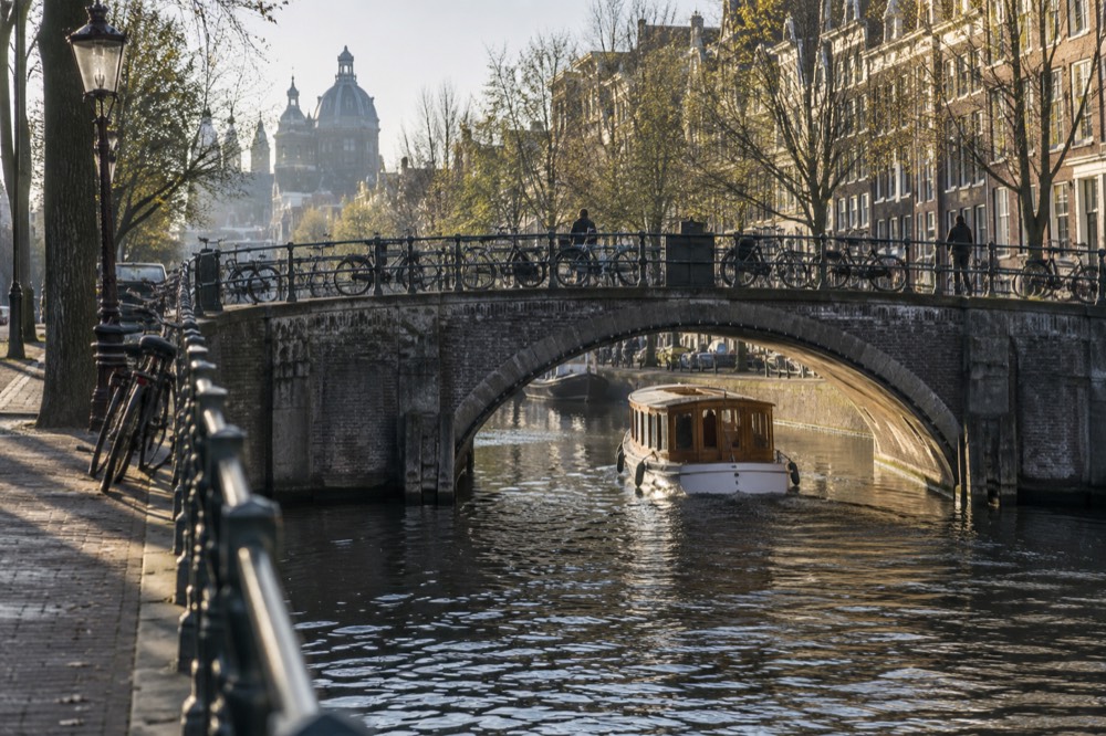 A small saloon boat passing under a stone canal bridge lined with bicycles